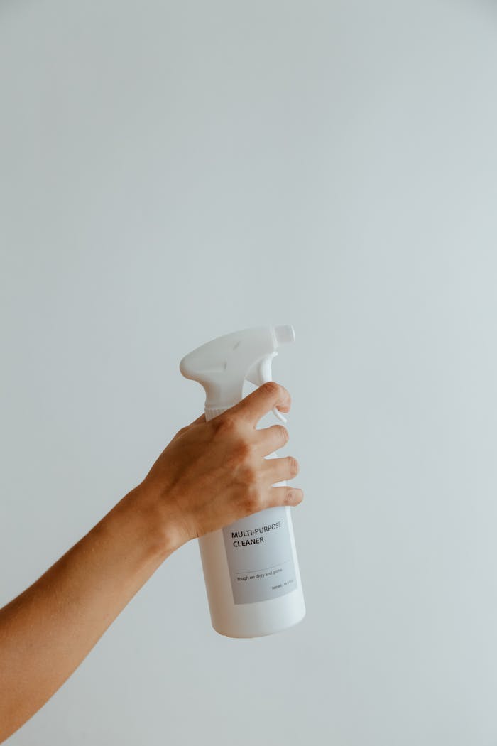 services-03 Close-up of a hand holding a white multipurpose cleaner spray bottle against a minimalistic background.