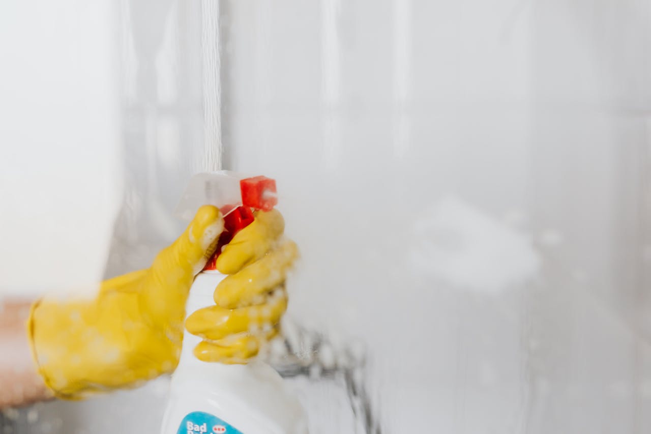 services-01 Crop unrecognizable person in yellow latex protective glove spraying detergent from bottle with plastic dispenser at glass of shower cabin
